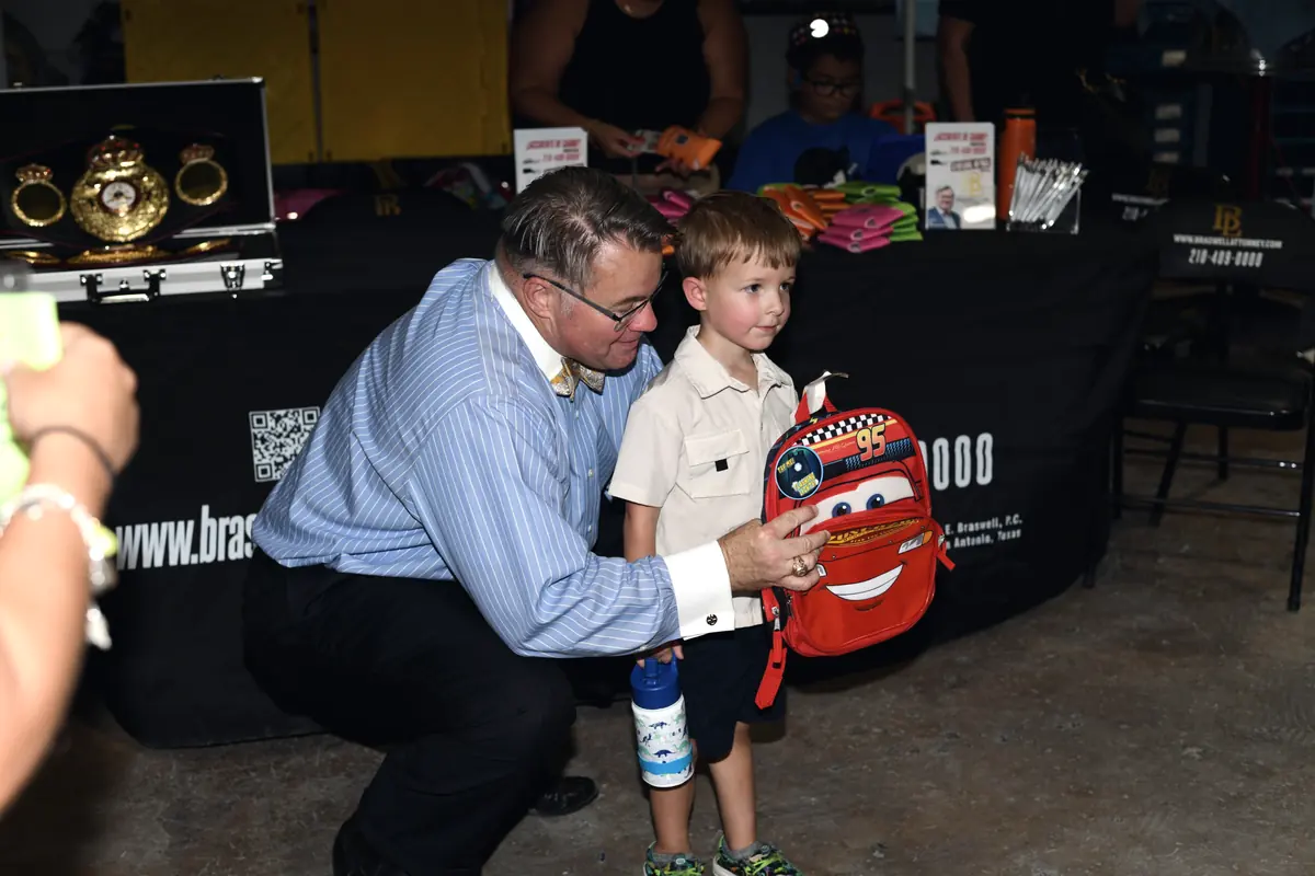 Mark Braswell kneeling to hand a Lightning McQueen backpack to a young boy at the Braswell Law tent