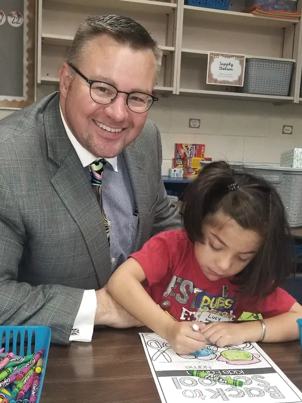 Mark Braswell sitting with a young student who is coloring a back-to-school worksheet
