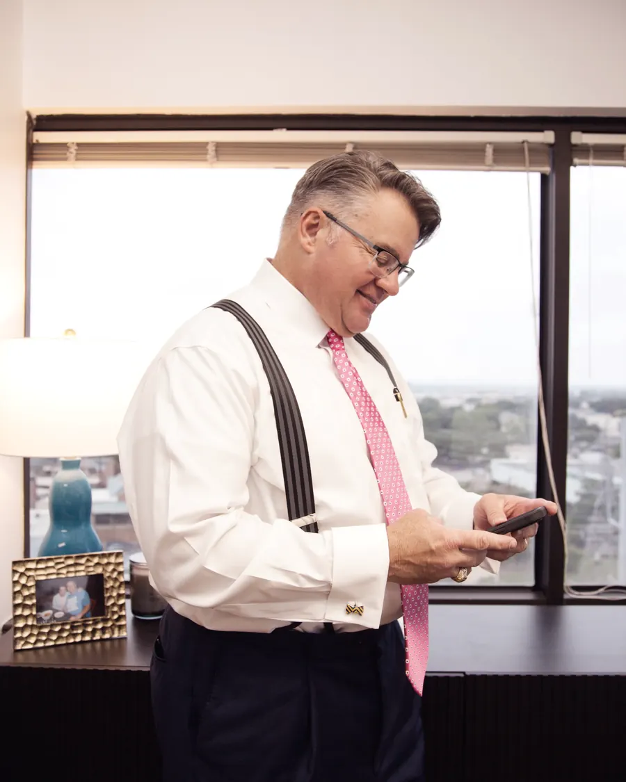 Mark Braswell standing at his office window, reading a message on his phone, with San Antonio behind him